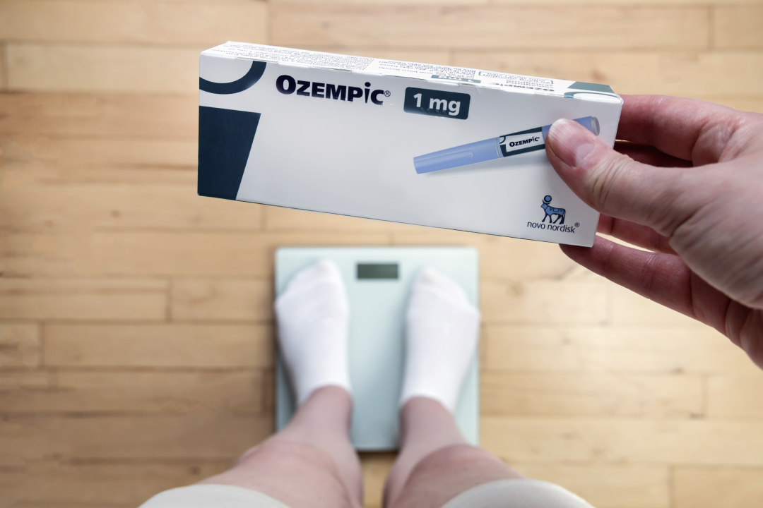 A person holds Semaglutide (Ozempic) while standing on a scale to track weight loss progress.