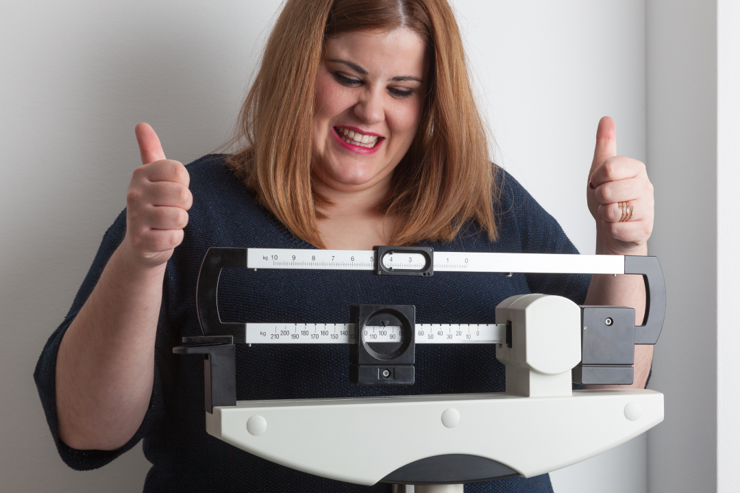 A woman smiling and giving thumbs up while standing on a scale, celebrating weight loss progress with Semaglutide.