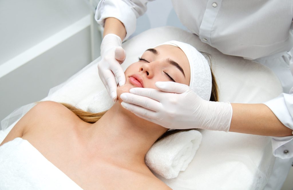 A woman lies on a treatment table with eyes closed as an esthetician wearing gloves holds her chin.