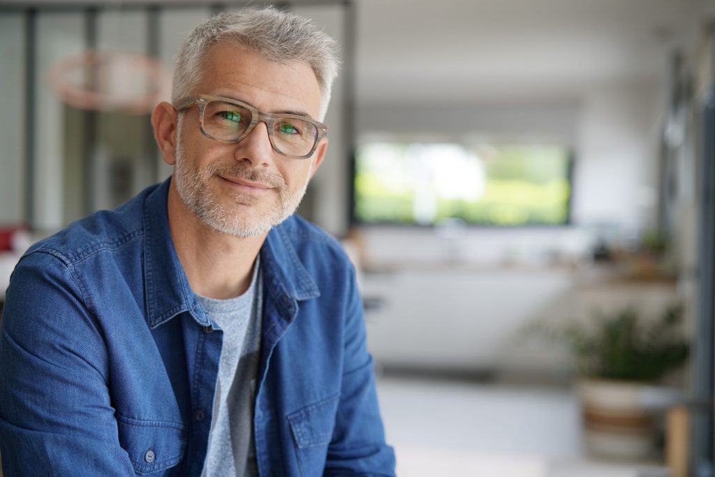 A middle-aged man with gray hair and glasses sits indoors wearing a denim shirt and looking toward the camera.