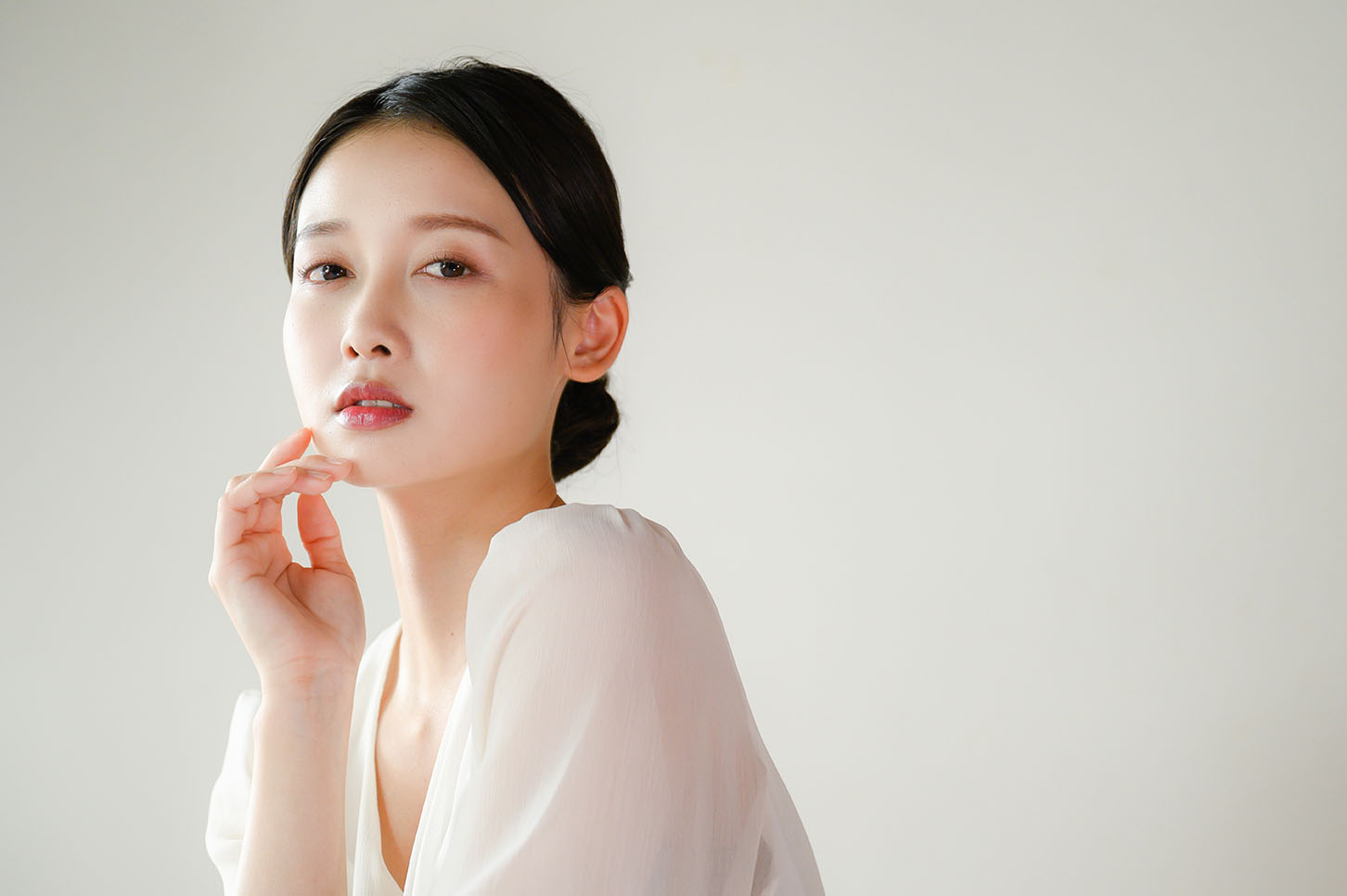woman in a white top looking toward the camera with one hand touching her chin against a neutral background